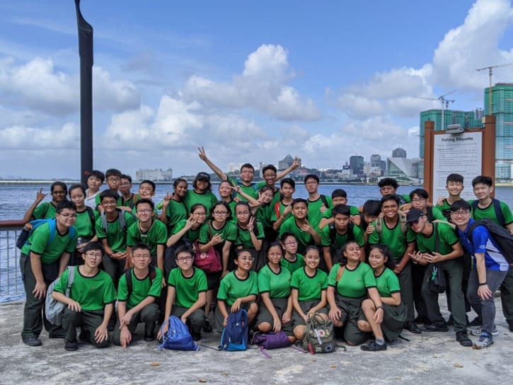Large group of youth in matching green shirts posing outdoors near a waterfront with a city skyline behind them.